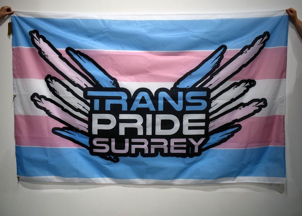 A blue, white and pink trans pride flag with "TRANS PRIDE SURREY" with 'wings' also in the trans pride colors.