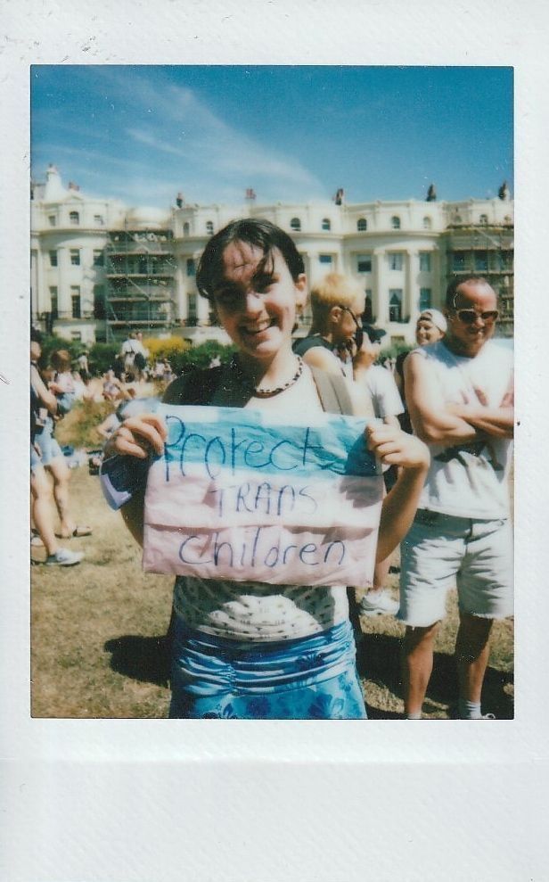 A person smiles while holding a sign reading "Protect Trans Children" at an outdoor gathering.