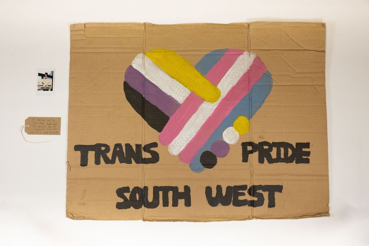 Hand painted brown cardboard placard with a stylised heart coloured in trans and non-binary colours and text "Trans Pride South West"