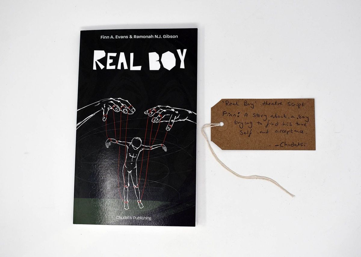 A black book which is called 'Real Boy' and has an illustration of a person being moved like a puppet. There is also a brown luggage tag with writing on it.