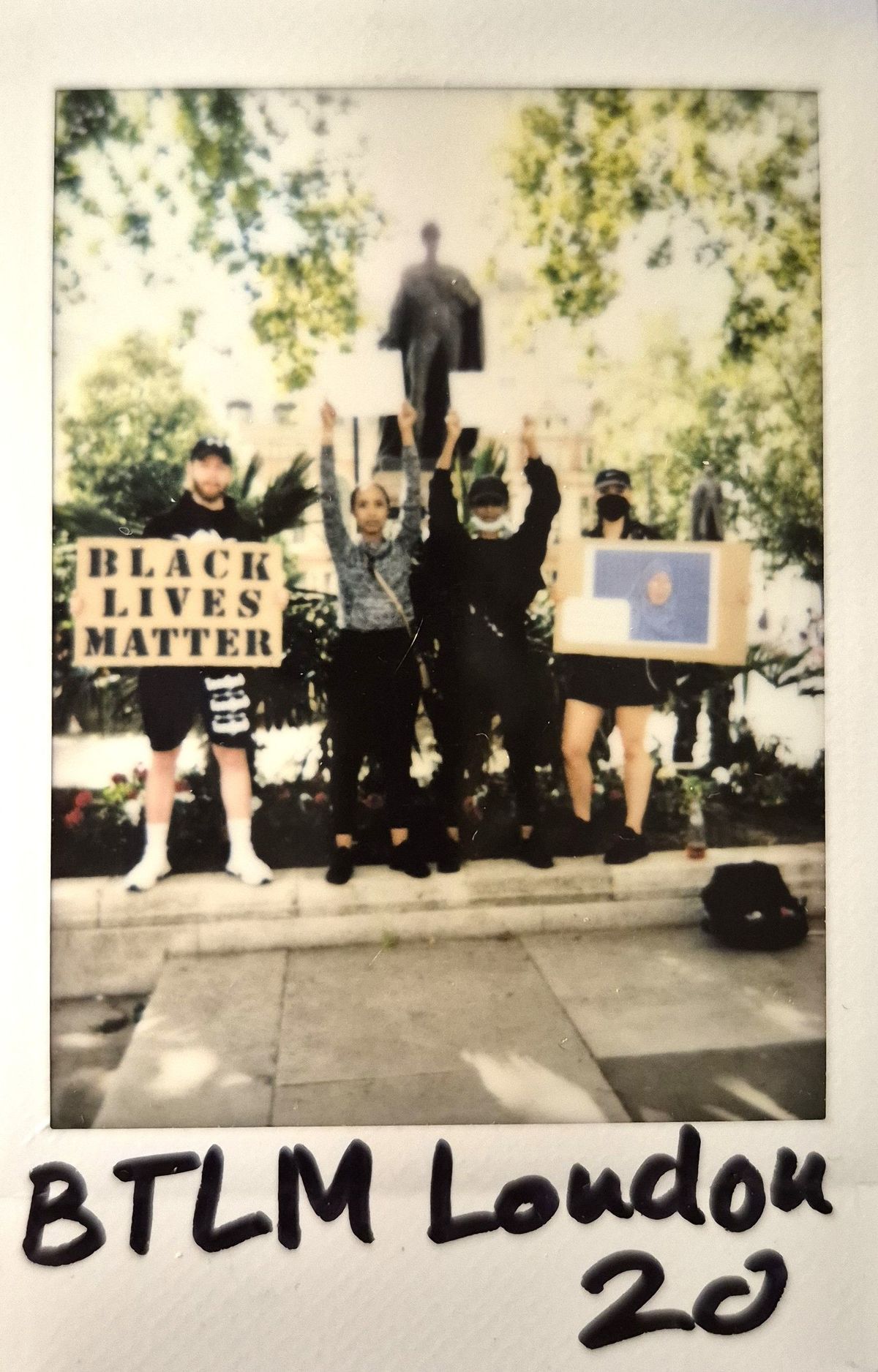 A diverse group stands with protest signs, supporting the Black Lives Matter movement, in a sunny park setting.