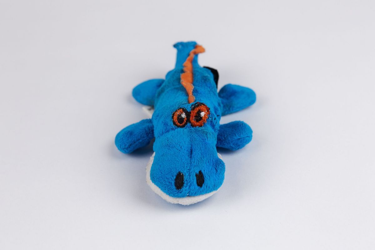 Blue, orange, and white crocodile-shaped dog toy. The eyes and spine of the toy have been visibly repaired with various threads.