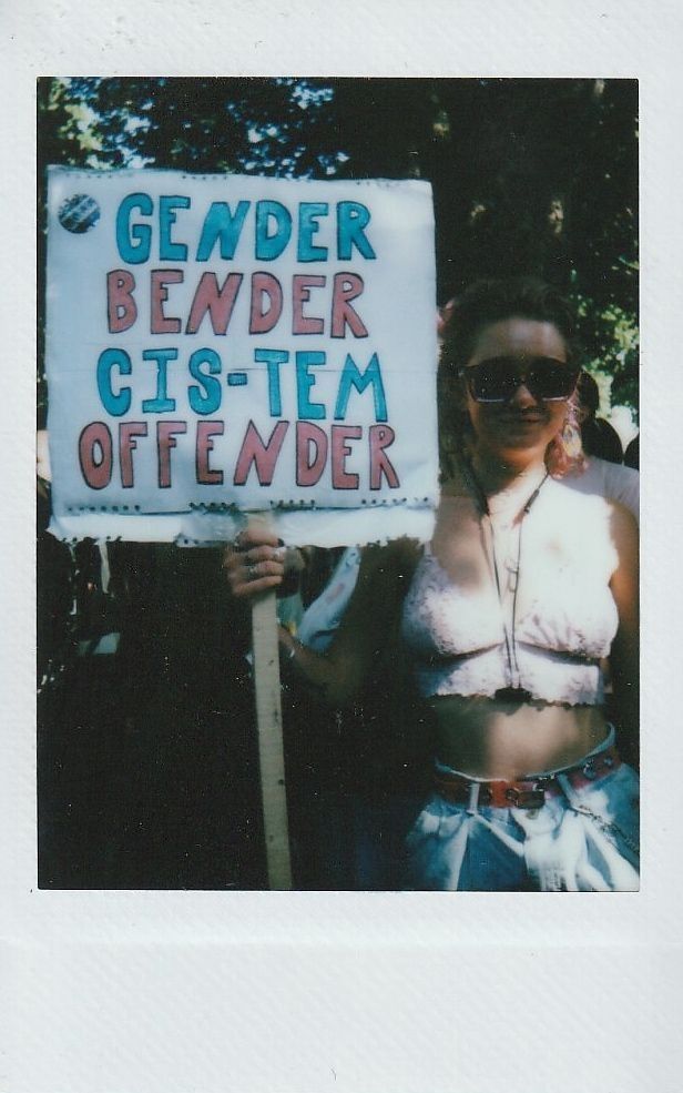 A person holding a sign reads "Gender Bender Cis-tem Offender" at an outdoor event.