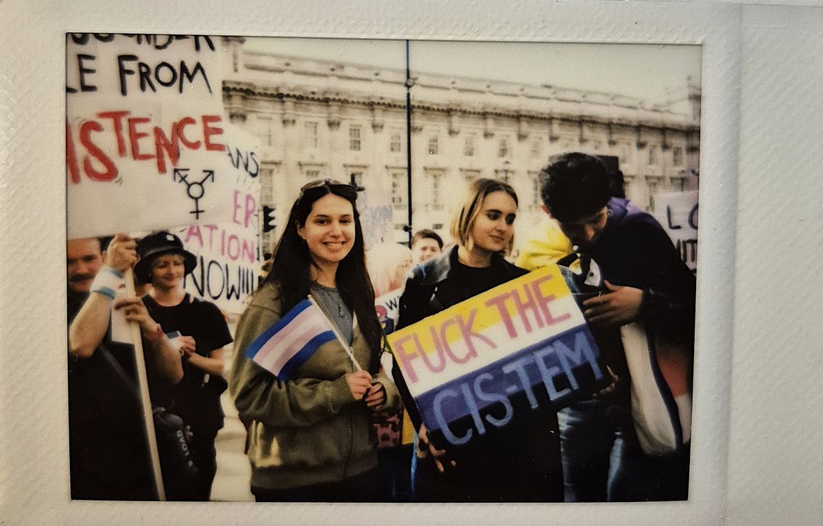 A group of people are standing at a protest, holding signs and smiling together in solidarity. The signs say 'FUCK THE CIS-TEM" and "RESISTANCE".