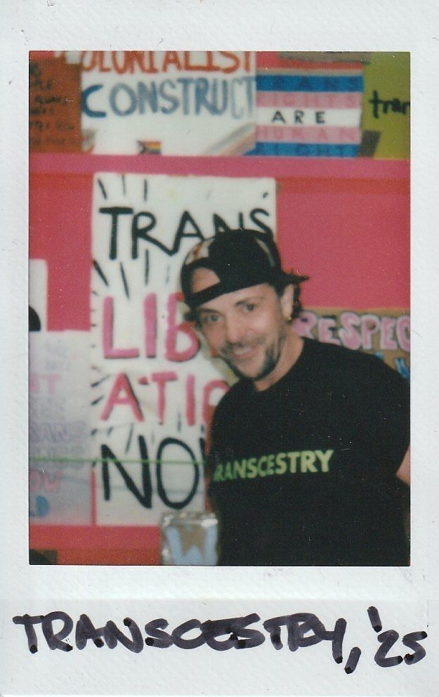 A person wearing a "TRANSCESTRY" shirt stands smiling in front of colorful posters.