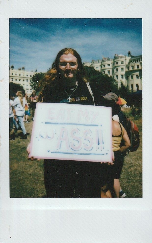 A person with long hair is holding a colourful sign which says 'EAT MY ASS".