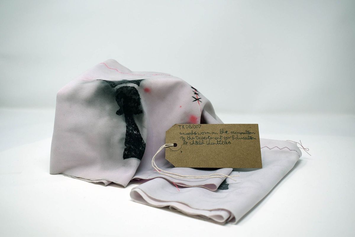 Two pale pink snoods decorated with the Trans Kids Deserve Better 'Pawn with a crown' logo. With a brown, handwritten luggage tag.