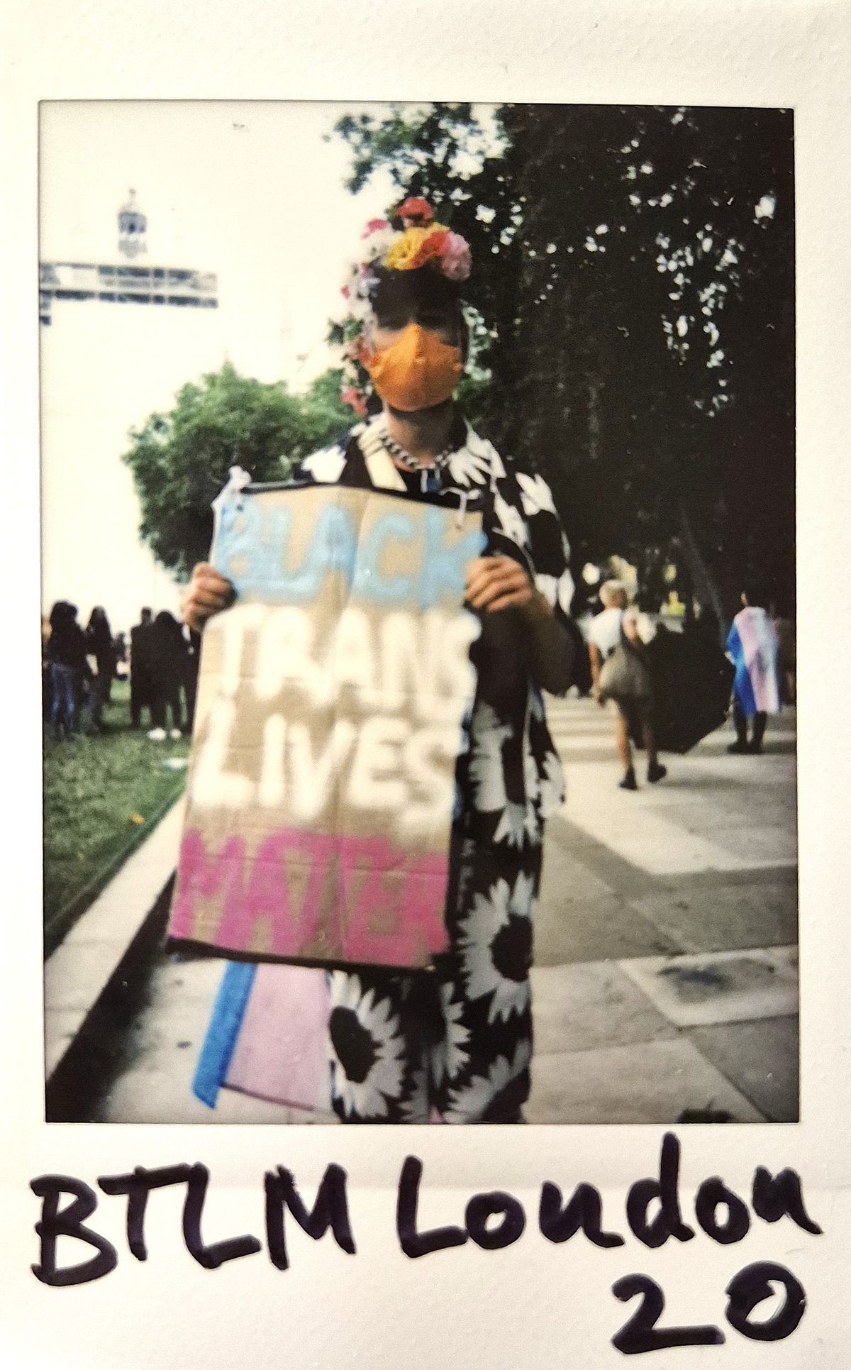 A person wearing a colorful mask and floral outfit holds a sign reading "Black Trans Lives Matter".