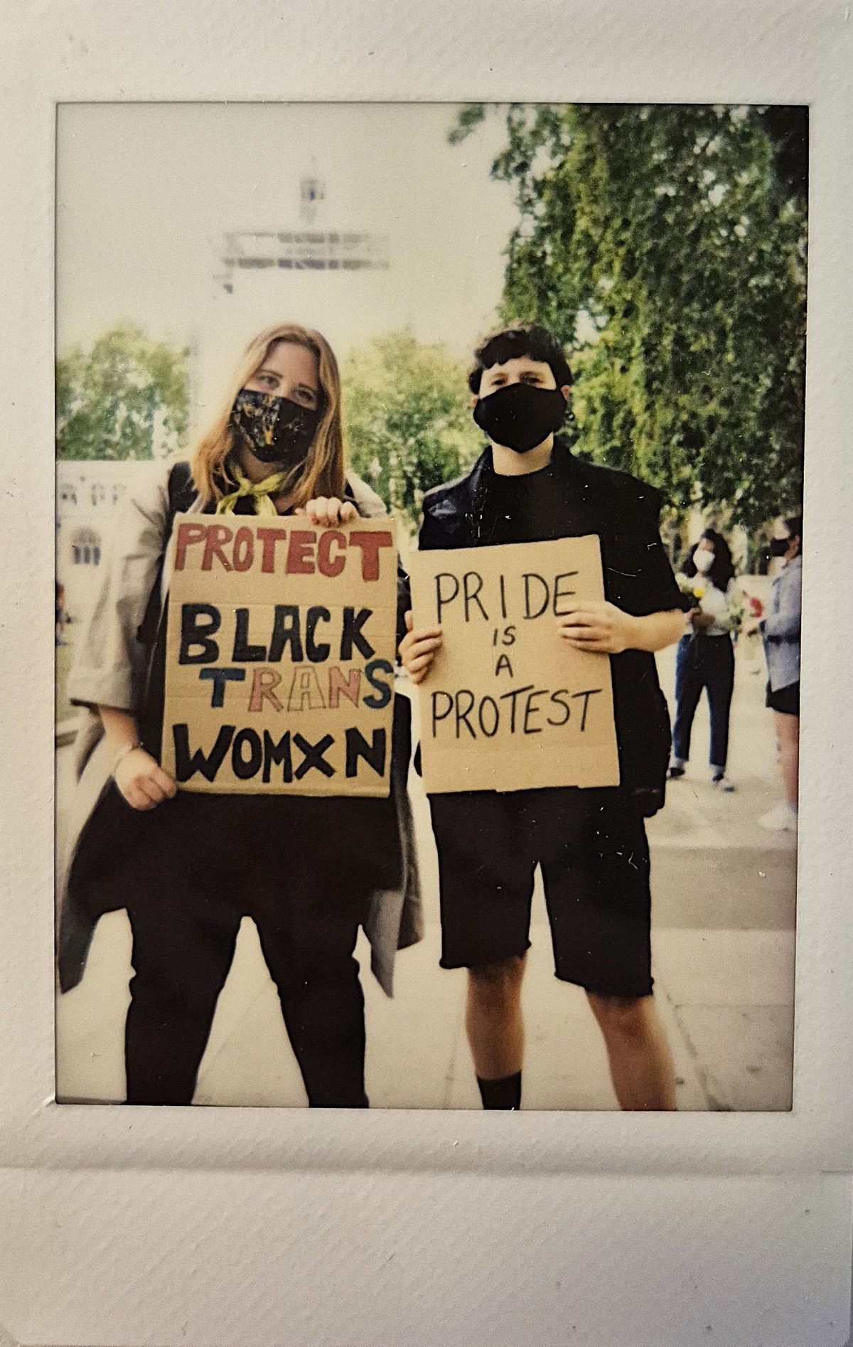Two masked individuals hold signs which say "PROTECT BLACK TRANS WOMXN" and "PRIDE IS A PROTEST".
