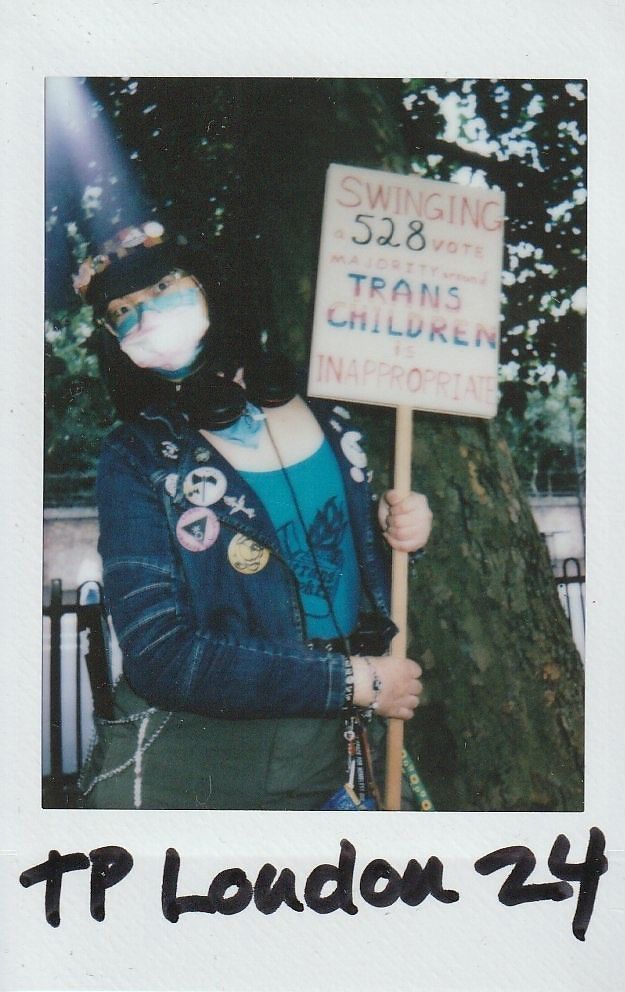 A masked person stands holding a sign expressing support for trans children under a tree.