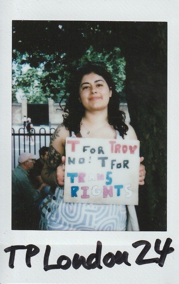 A person stands under a tree holding a sign advocating for trans rights in colorful letters.