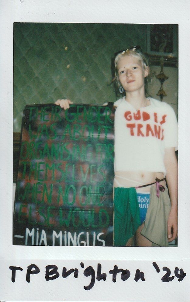 A person stands holding a colorful sign with a quote, wearing a shirt with "God is Trans."