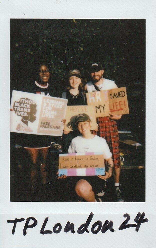 Four people are posing together outdoors, holding various protest signs that express support for trans rights.