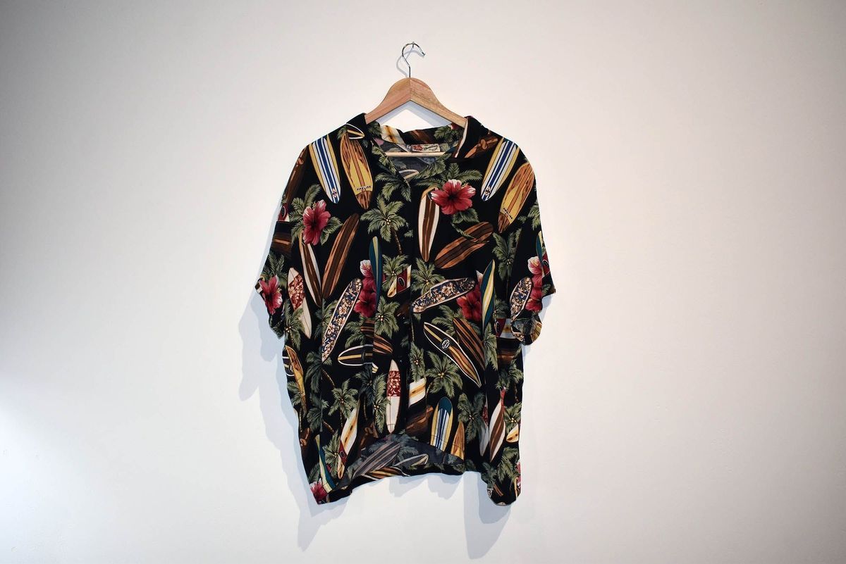 A colourful Hawaiian shirt featuring surf boards, green leaves and red flowers on a back background, on a wooden hanger.