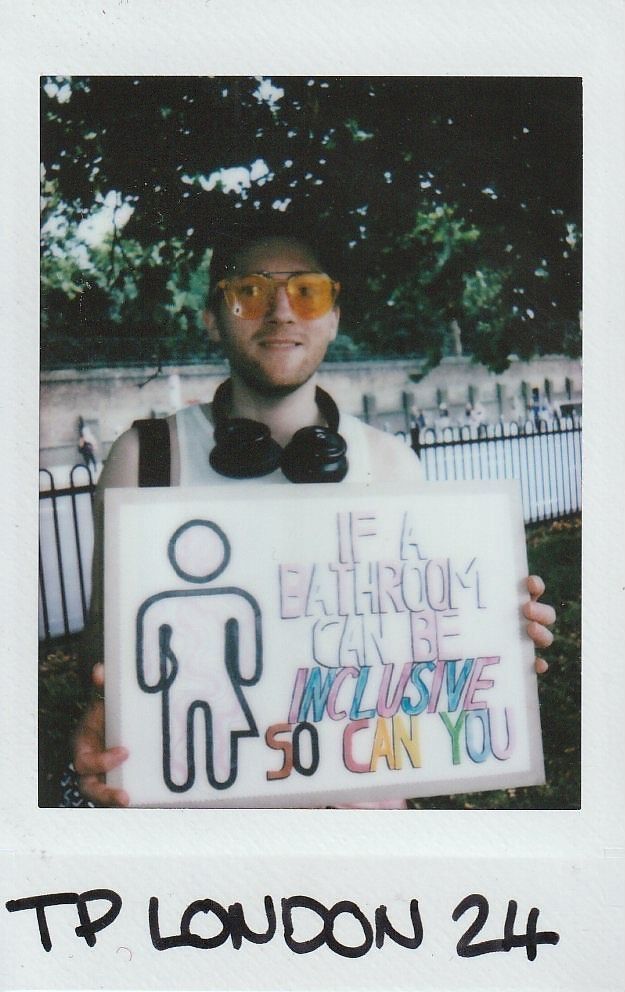 A person holds a sign saying, "If a bathroom can be inclusive, so can you," outdoors.