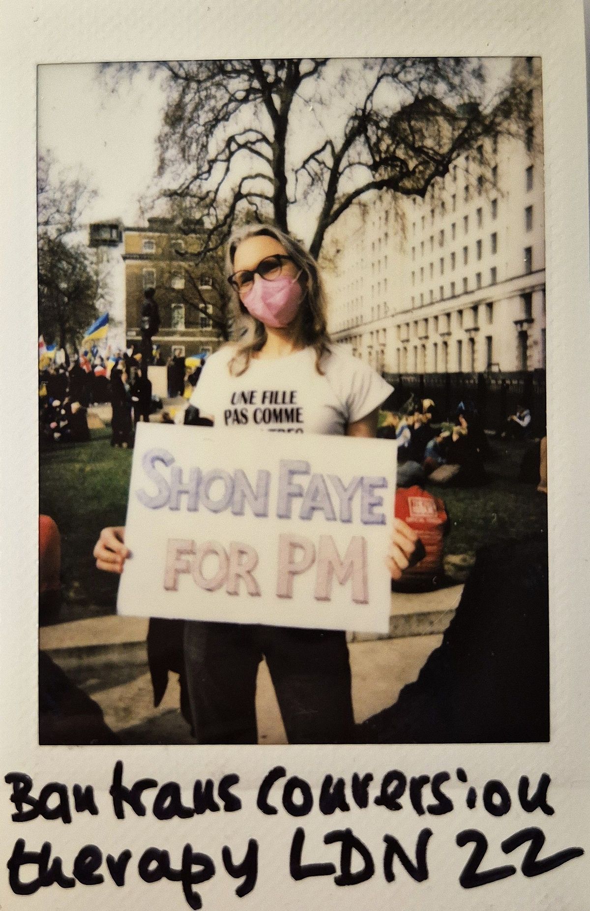 A person stands outdoors holding a sign reading "Shon Faye for PM," wearing a pink mask.