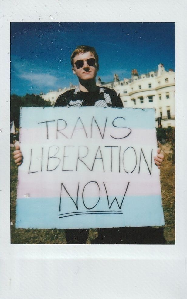 A person holds a sign saying "Trans Liberation Now," standing outside near a white building.