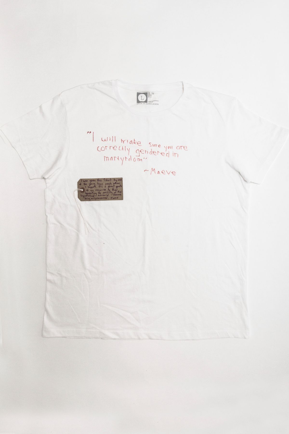 A white T-shirt features a quote in red handwriting and a small handwritten cardboard tag.