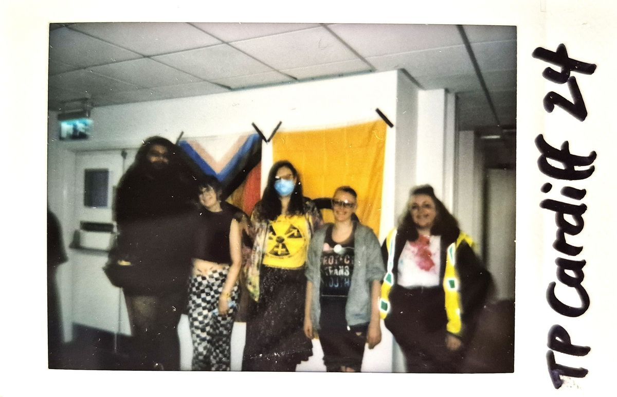 Five people pose together in front of colorful flags on a wall, smiling cheerfully indoors.