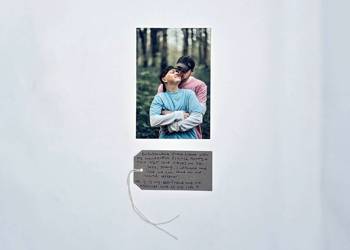 A photo of two people tenderly embracing in a wood celebrating their engagement. Both wear baseball caps, one a blue t-shirt, the other a pink t-shirt. With a brown handwritten luggage tag.