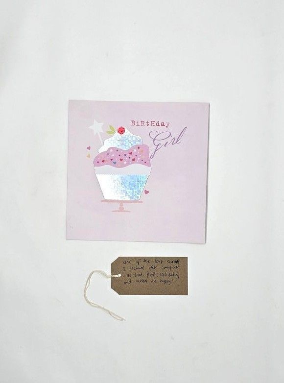 A Birthday Card which says 'Birthday Girl' and has an illustration of cupcake. The card is predominantly pink colours. Underneath the greeting card is a brown tag with writing on it.