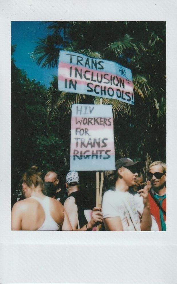A group of people hold signs, one which says "TRANS INCLUSION IN SCHOOLS" and the other "HIV WORKERS FOR TRANS RIGHTS".