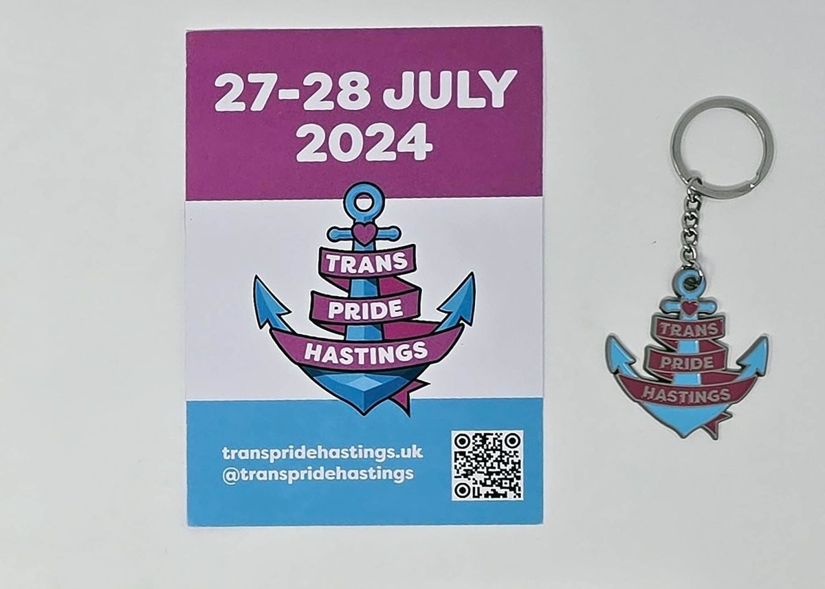 A blue, white and pink flyer for Trans Pride Hastings 2024 and an enameled metal keyring in the shape of the Trans Pride Hastings logo. The logo is a blue anchor with a pink heart a the top and the words "Trans Pride Hastings" in white on a pink ribbon entwined round the anchor.