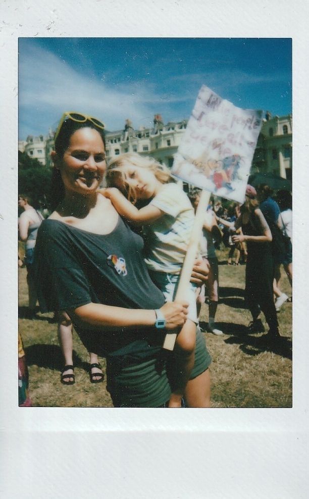 A smiling woman holds a sign with a child at an outdoor gathering on a sunny day.