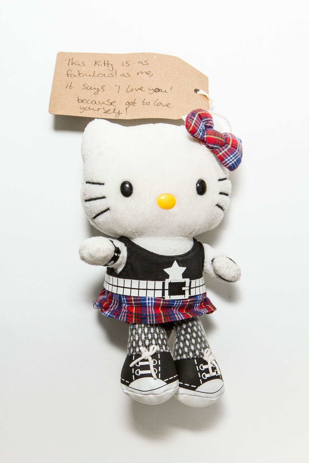 Soft Hello Kitty character toy wearing a plaid bow on one ear, black sleeveless shirt, studded belt, plaid skirt, leggings, wrist cuffs, and black and white shoes.