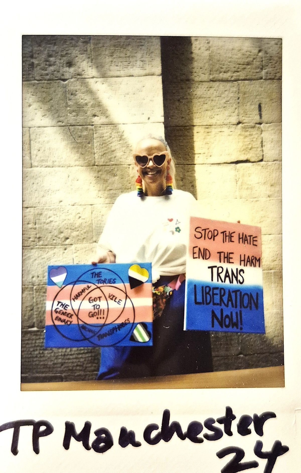 A person with heart sunglasses holds signs advocating for trans liberation against a brick wall backdrop.