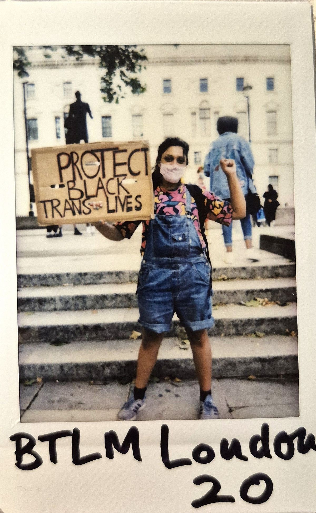A person wearing a mask holds a sign saying "Protect Black Trans Lives," standing on steps.