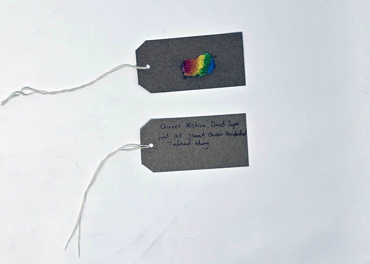 A piece of rainbow colour duct tape stuck to a brown luggage tag. With a second brown, handwritten luggage tag.