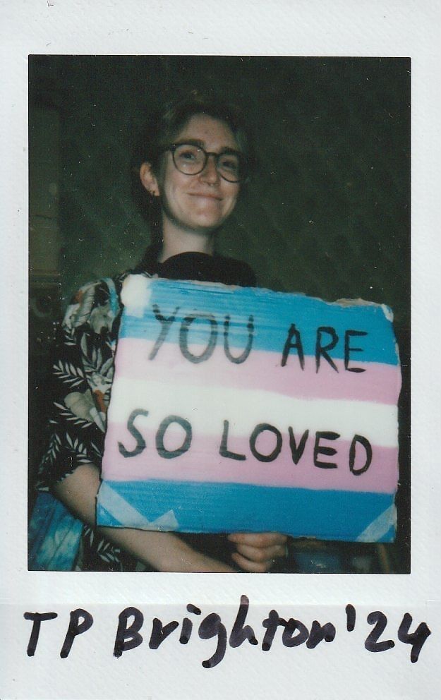 A person smiles while holding a colorful sign that reads, "You Are So Loved" in black letters.