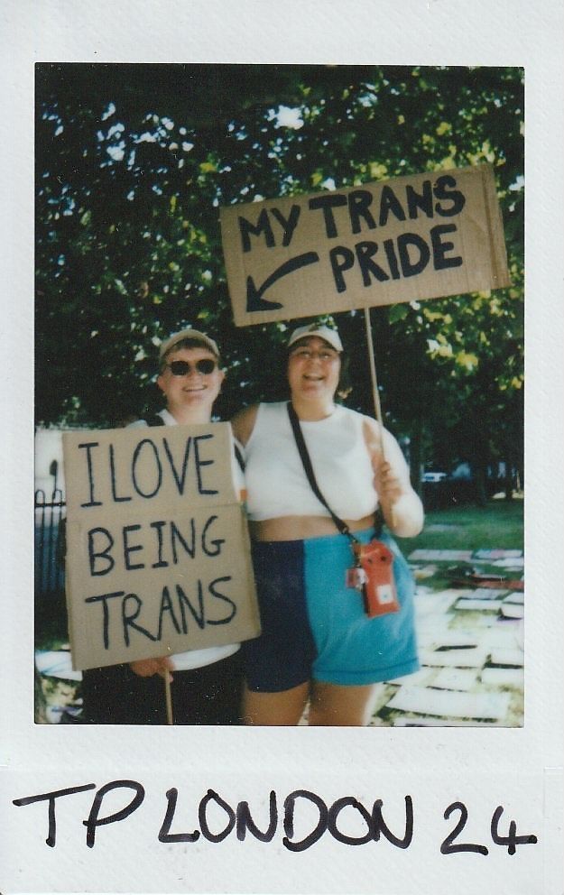 Two people are holding signs that express their pride and love for being transgender at an outdoor event.