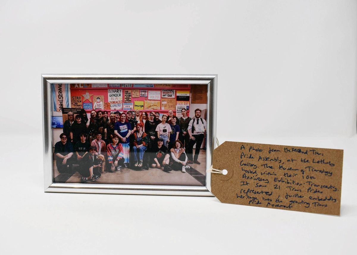 The image shows a group photo in a silver frame, with colourful protest signs hung on a pink wall at an exhibition. Next to the framed photograph is a brown tag with handwritten text on it.