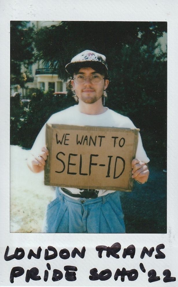A person in glasses holds a cardboard sign saying “We want to self-ID” at an event.