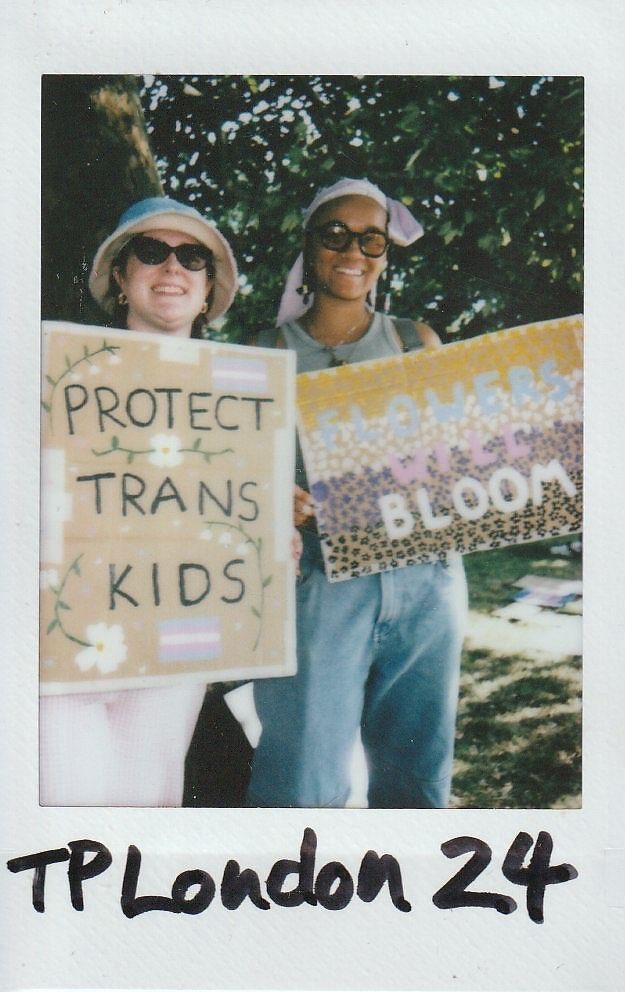 Two individuals are smiling and holding signs with supportive messages about trans rights, standing outdoors.