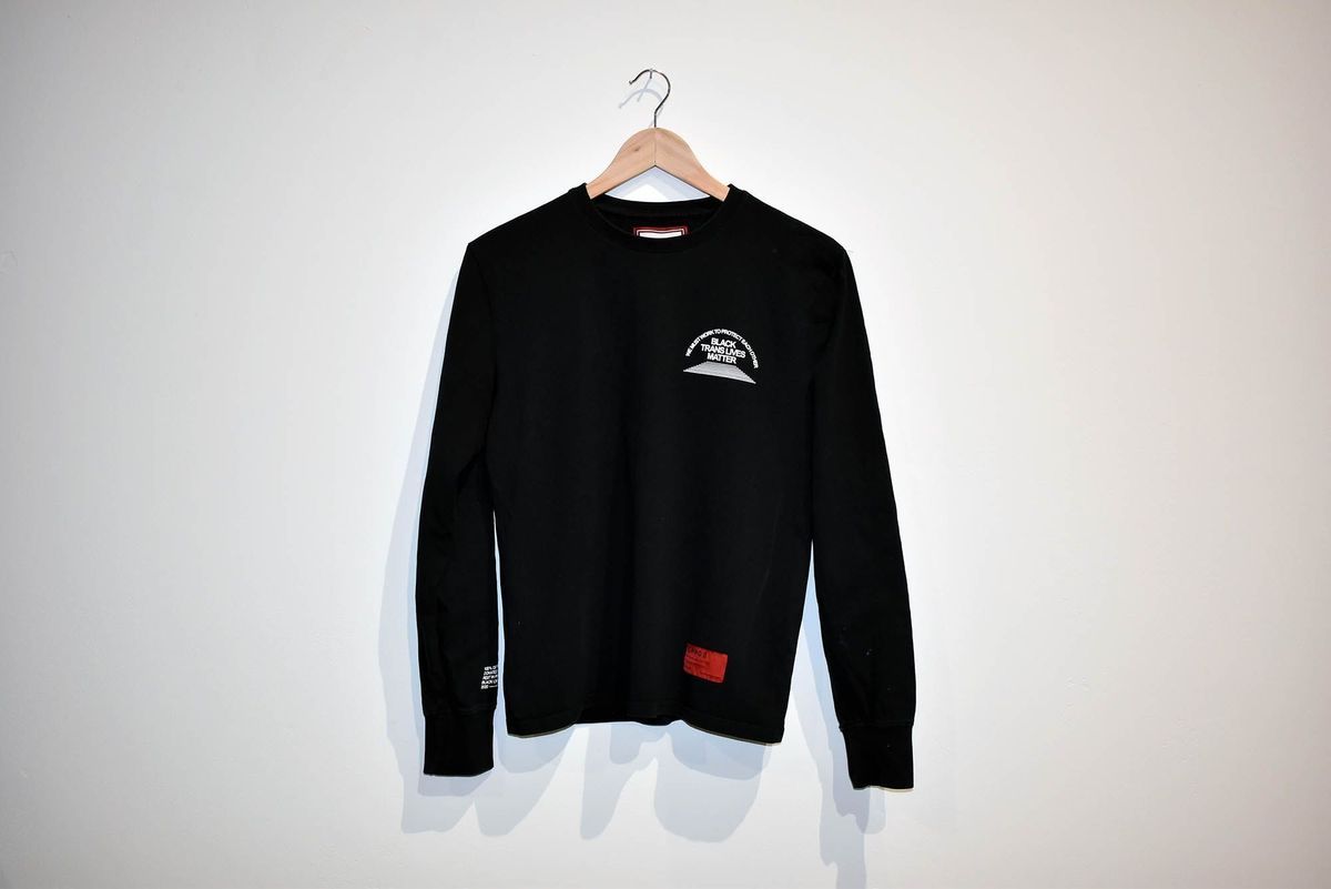 A black long-sleeve shirt is hanging on a wooden hanger against a plain wall. On the shirt, it says 'We Must Work To Protect Each Other' and 'Black Trans Lives Matter'.