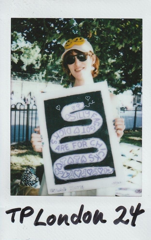 A person wearing sunglasses and a cap holds a colorful sign with playful text at an outdoor event.