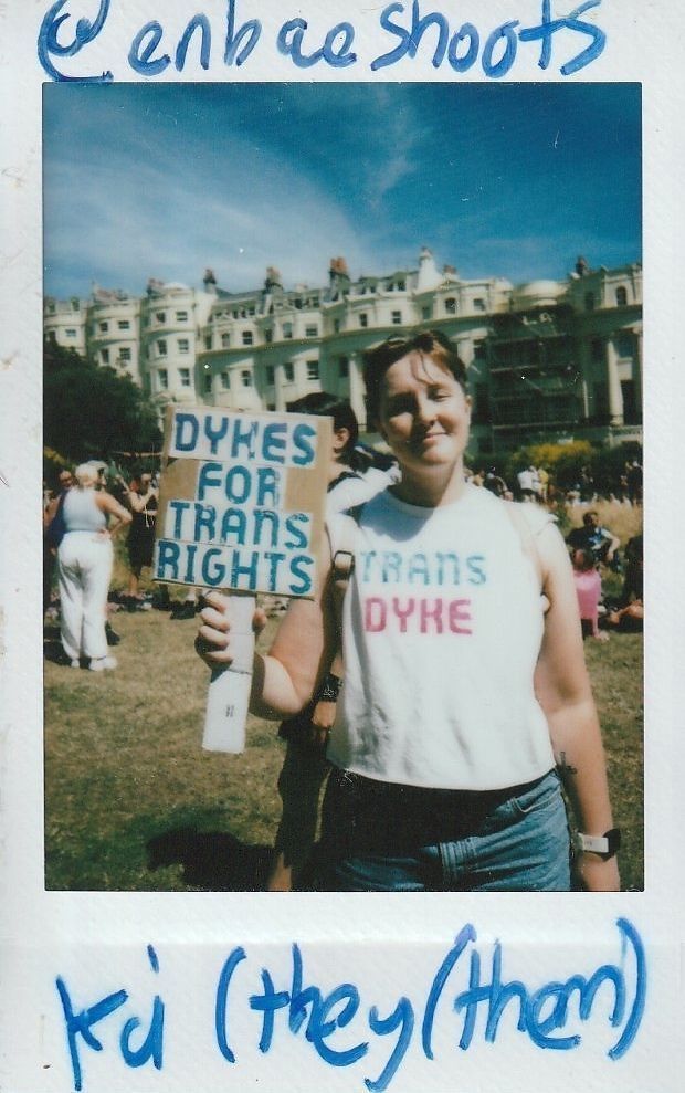 A person holds a sign reading "Dykes for Trans Rights," wearing a shirt with "Trans Dyke" printed on it.