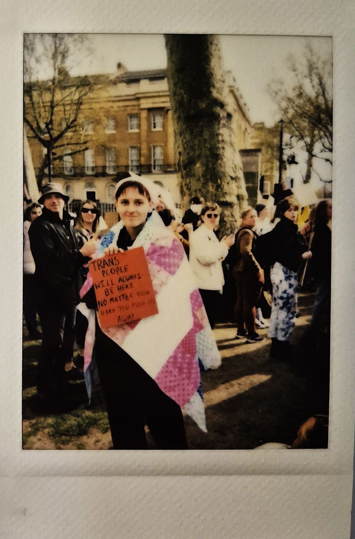 A person wrapped in a flag holds a sign at a gathering, surrounded by people and trees.