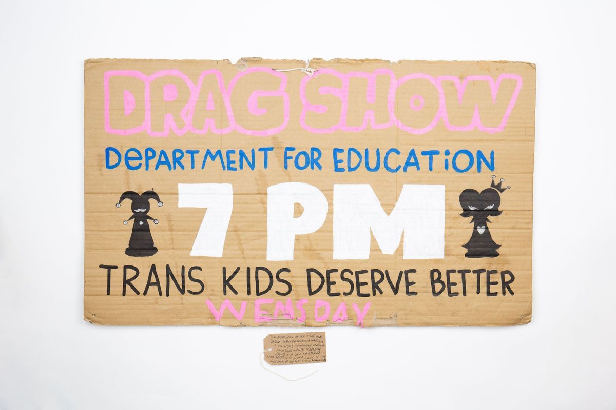 A hand drawn cardboard placard advertising a Drag Show a the Department for Education organised by Trans Kids Deserve Better. With a brown, handwritten luggage tag.