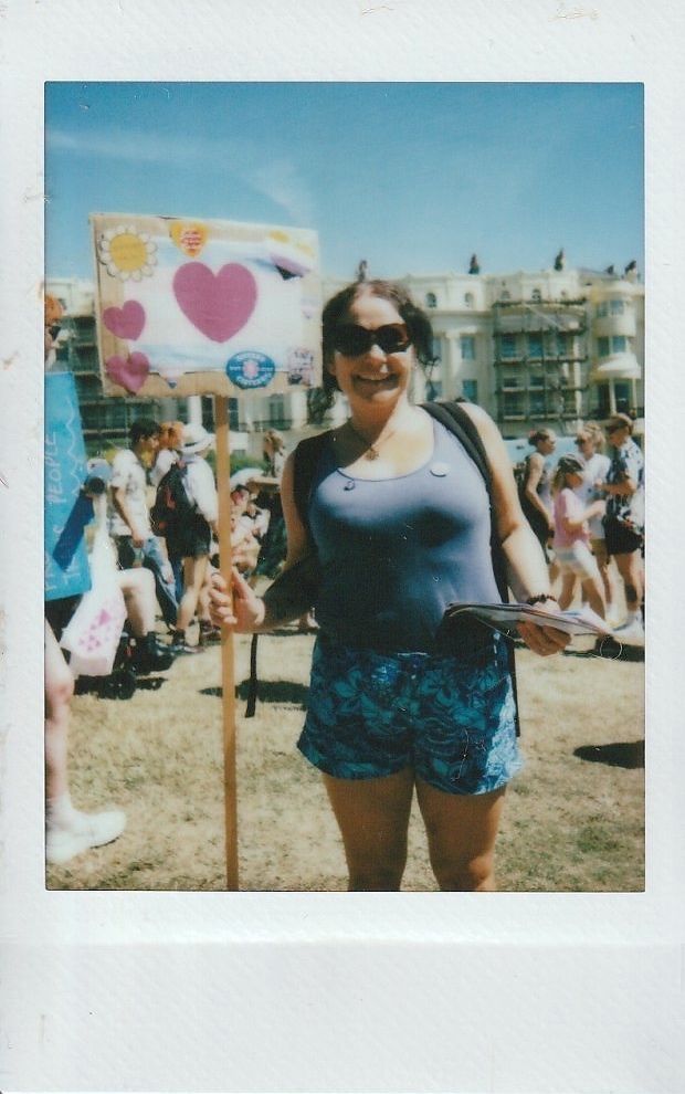 A smiling person holds a sign with hearts during an outdoor event on a sunny day.