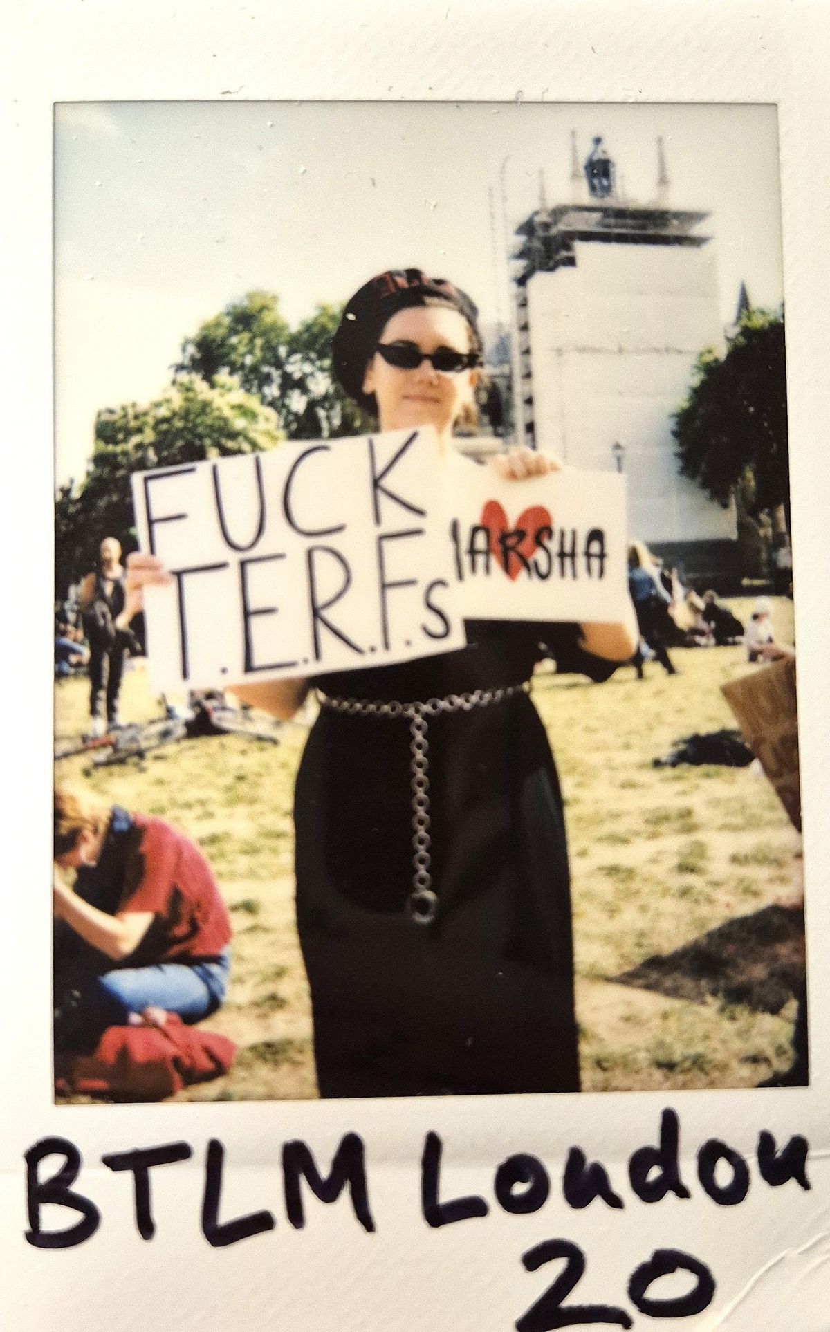 A person in sunglasses and a hat holds a sign which says 'FUCK TERFS' in a sunny outdoor setting.