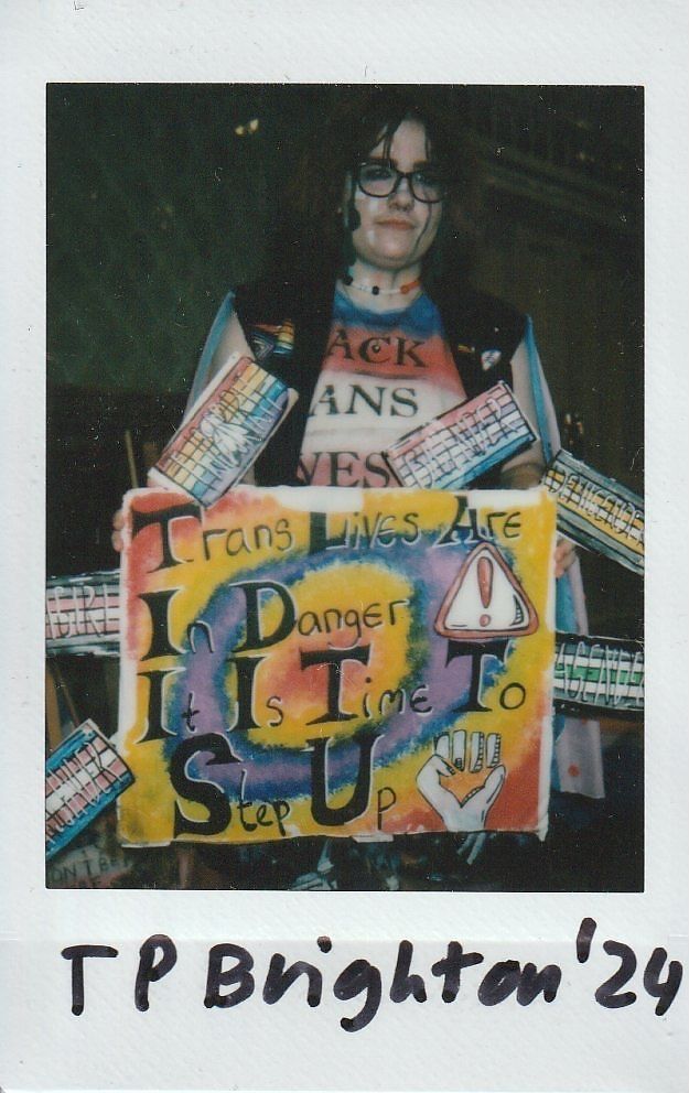 A person holds a colorful protest sign advocating for trans lives and action, with various badges displayed.
