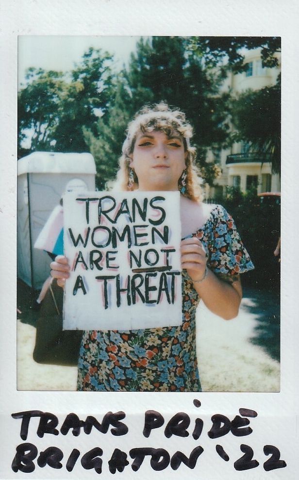 A person holds a sign reading "Trans women are not a threat" at an outdoor event.