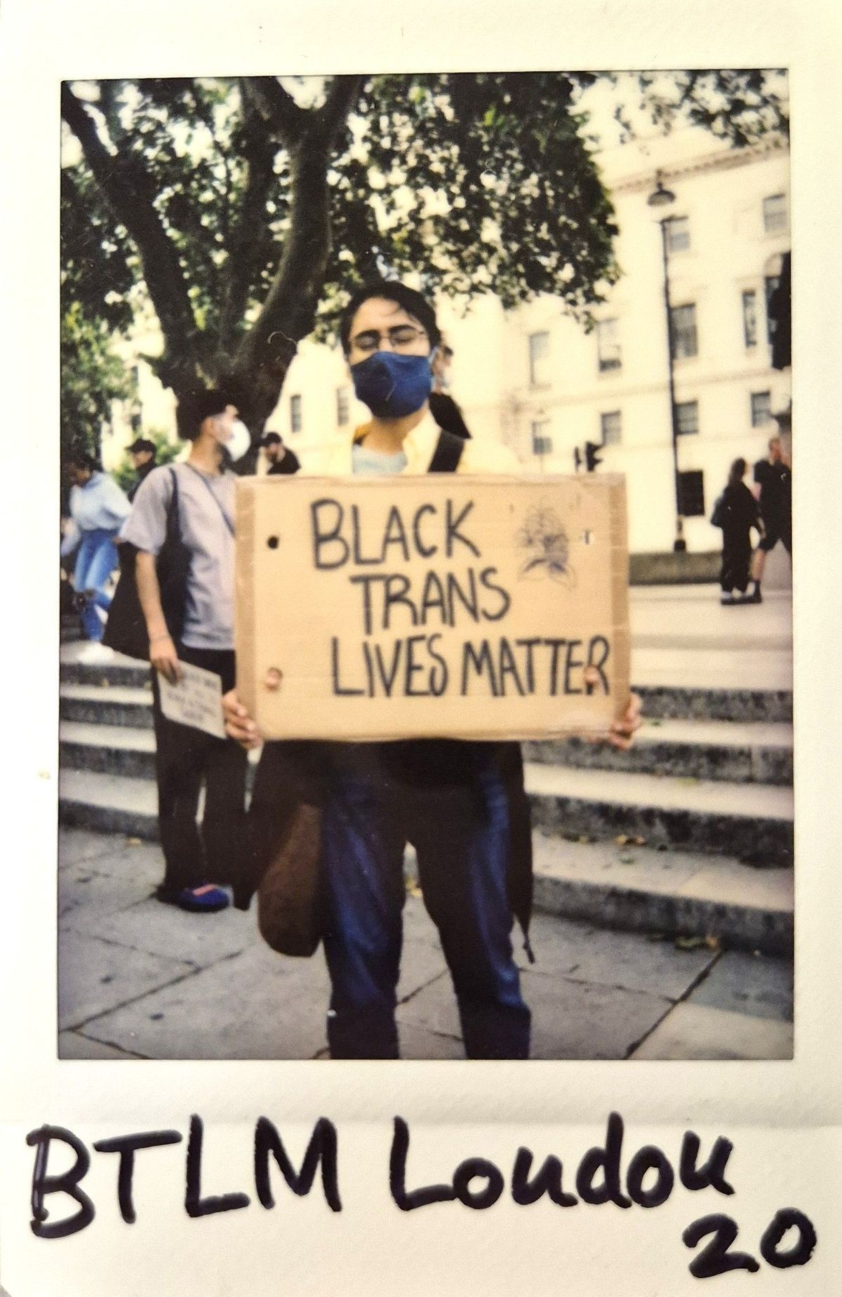 A masked individual holds a cardboard sign that reads "Black Trans Lives Matter" during a protest.