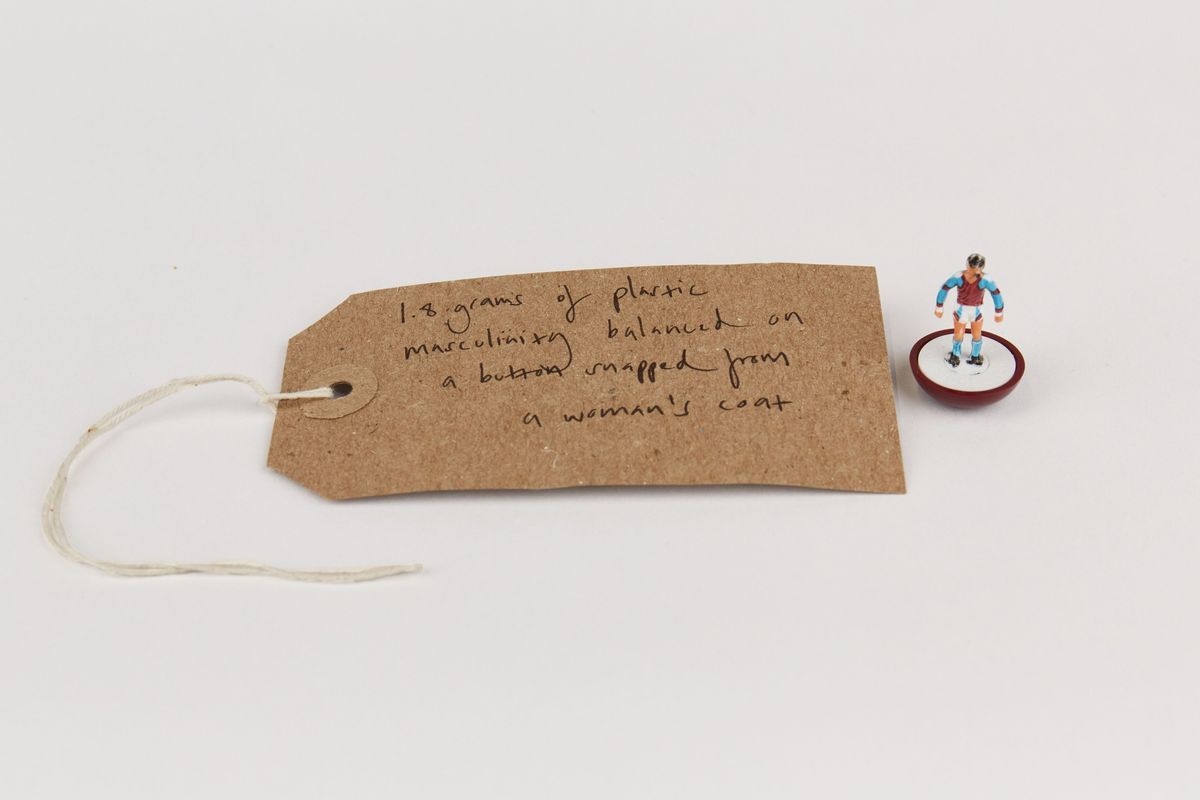 A small figure stands on a button beside a handwritten tag describing plastic masculinity weight.