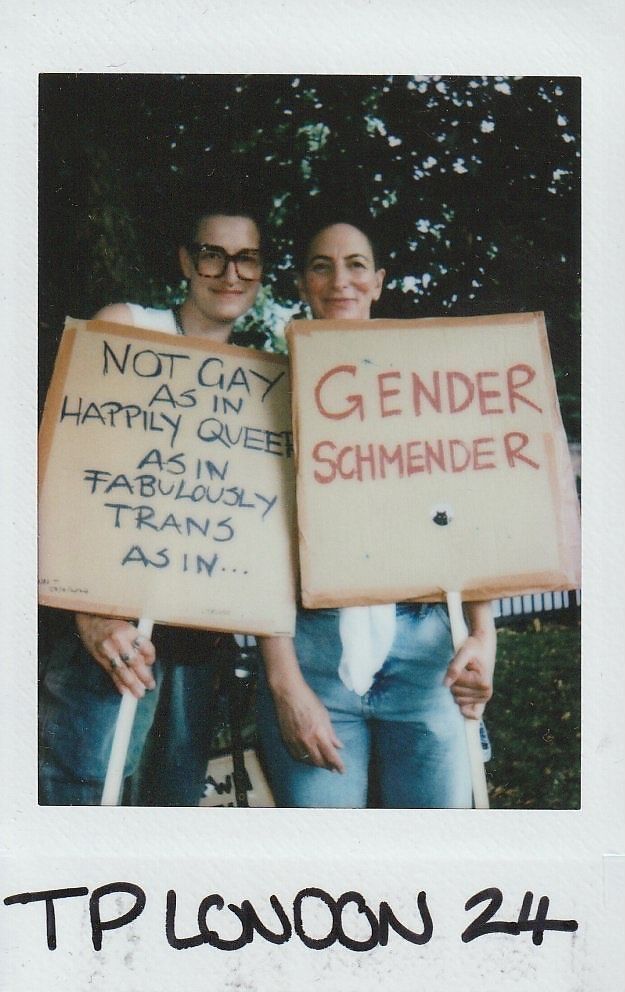 Two people hold signs with messages celebrating queer and transgender identities, standing confidently together outdoors.