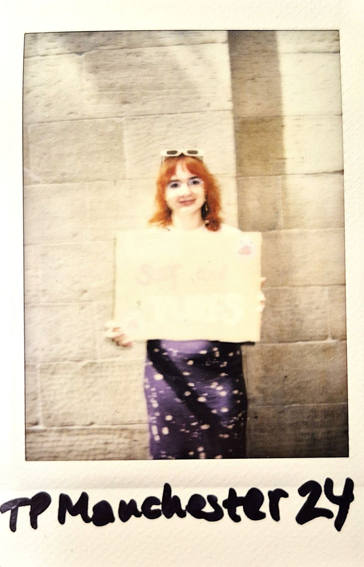 A polaroid of a person with red hair holds a partially visible sign while standing against a beige wall.